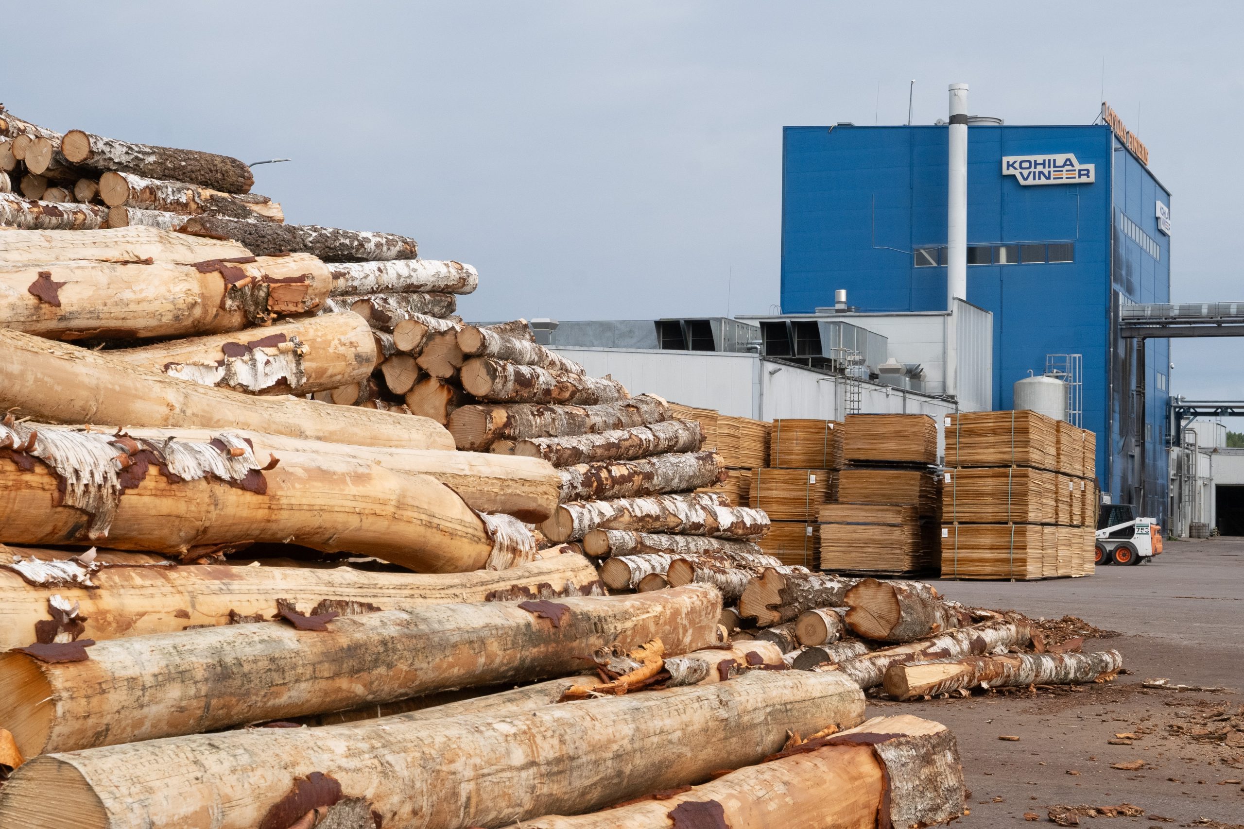 Blue plywood mill with the logo "Kohila Vineer" on it with logs and veneer besides it