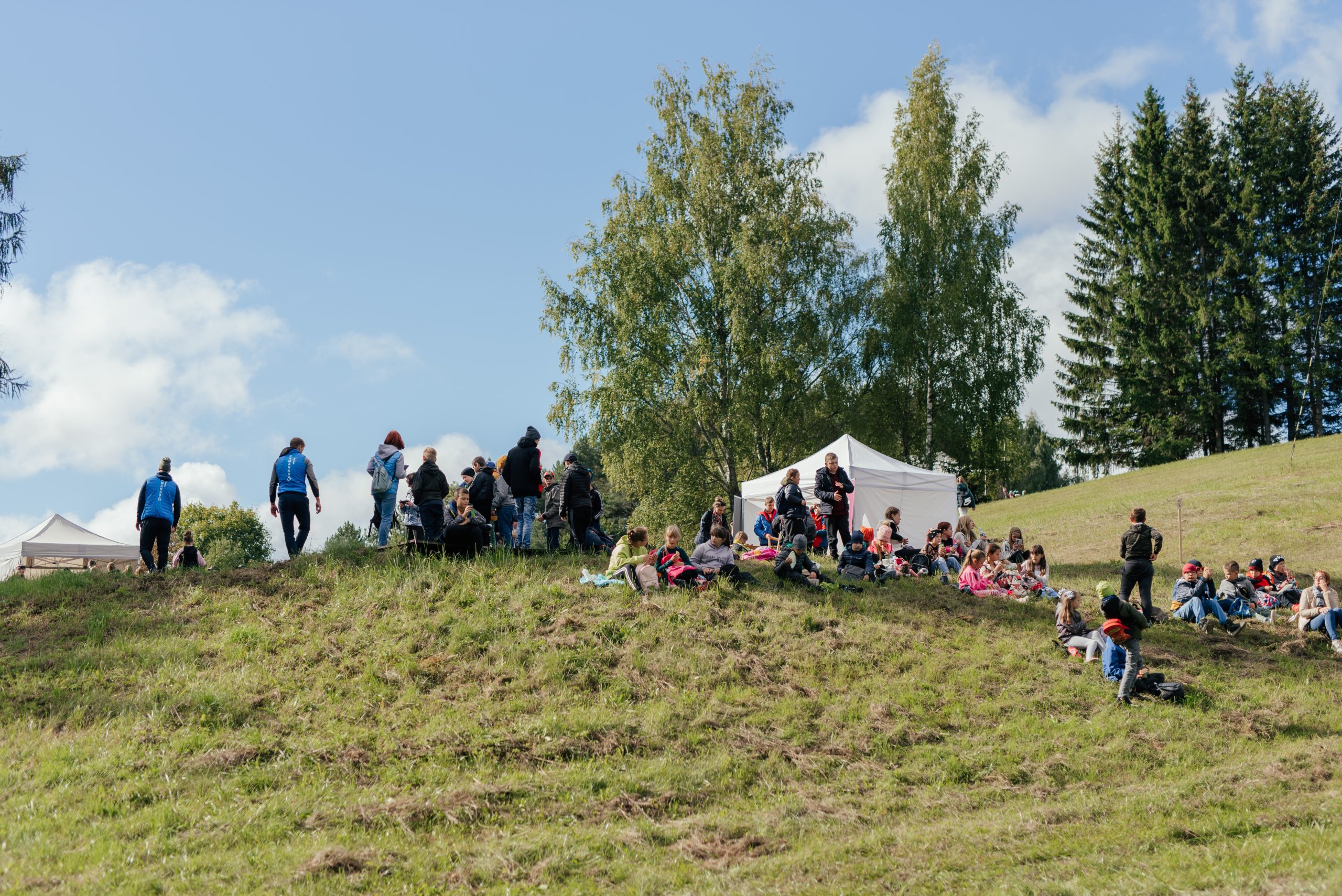 Bērni ar skolotājām sēž uz kalniņa netālu no kokiem