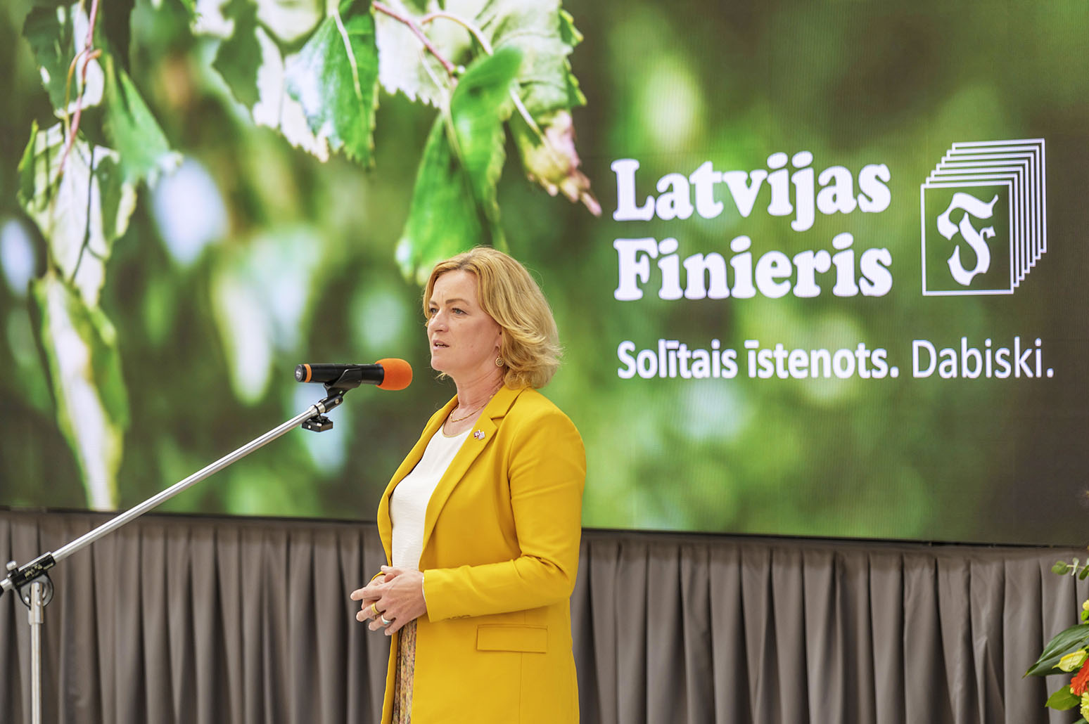 Sieviete runā mikrofonā ar lielu "Latvijas Finieris" logo uz ekrāna aizmugurē