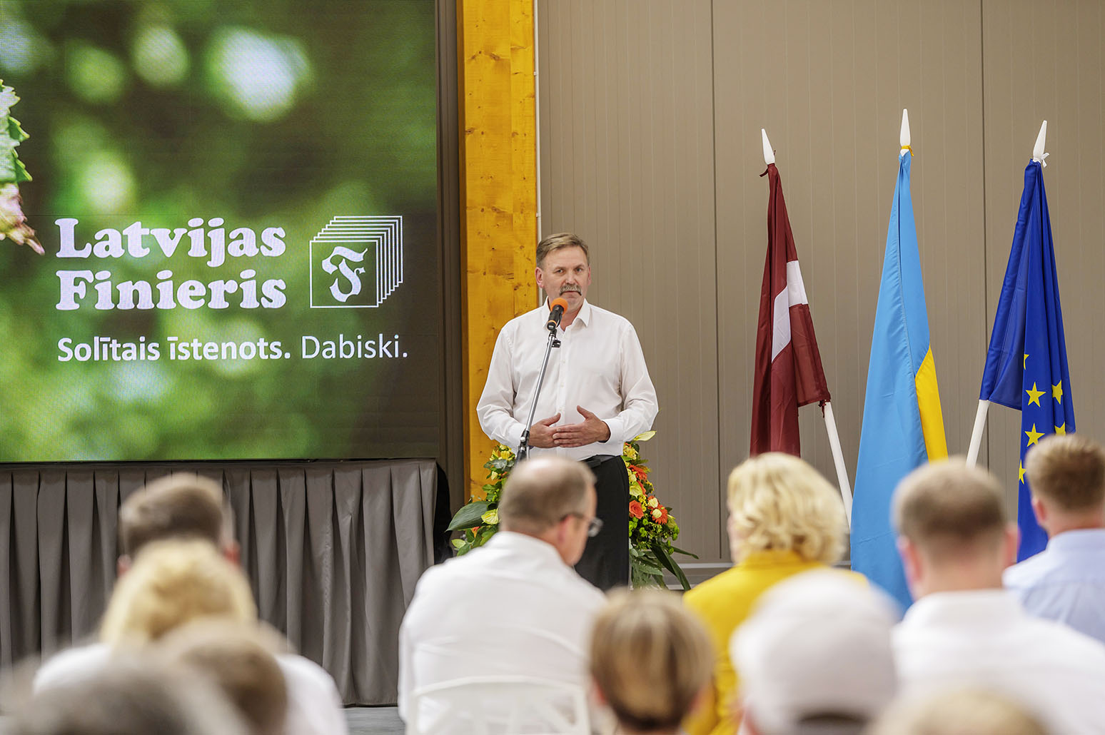 Vīrietis runā mikrofonā ar lielu "Latvijas Finieris" logo uz ekrāna aizmugurē